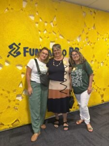 Viviani Kwecko (Coordenadora e-COO), Júlia Oliveira (Agricultora e-COO) e Lúcia Nobre (Coordenadora e-COO) em frende a fundo amarelo de tecido com logo e texto escrito "Fundação BB"