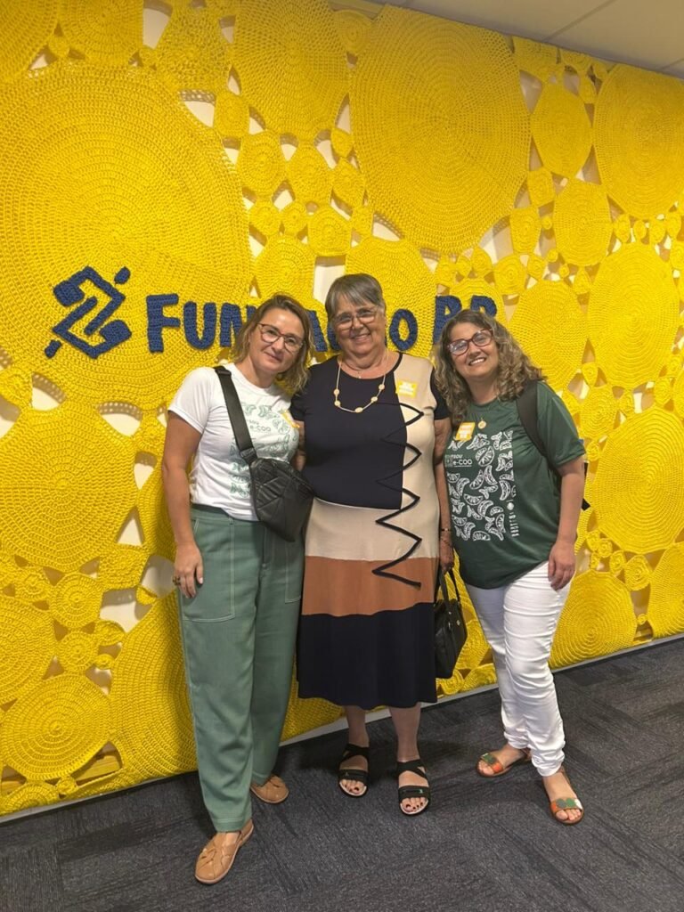 Viviani Kwecko (Coordenadora e-COO), Júlia Oliveira (Agricultora e-COO) e Lúcia Nobre (Coordenadora e-COO) em frende a fundo amarelo de tecido com logo e texto escrito "Fundação BB"